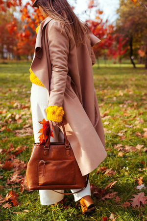 Fashionable woman wearing stylish clothes and accessories walking in fall park. Autumn female outfit. Trendy flared pants with yellow sweater, beige coat and brown handbagの写真素材