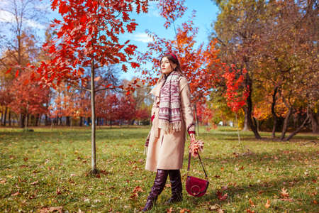 Full body portrait of beautiful woman wearing stylish warm clothes, boots, accessories in autumn park among red trees. Female fall fashion.の写真素材