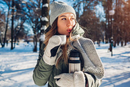 Woman drinking hot tea holding vacuum flask in winter park. Drinks to warm up in snowy frosty...