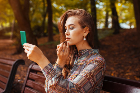 Portrait of young woman applying lipstick using hand mirror in fall park sitting on bench. Girl checking makeup outdoorsの写真素材