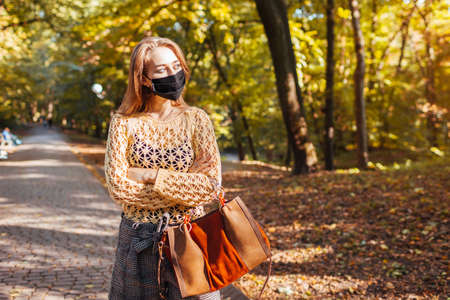 Pretty young woman wears protective mask walking in autumn park holding leaves.の写真素材