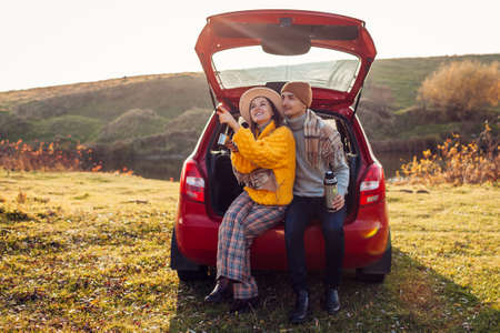 Loving couple relaxing in car trunk by autumn lake drinking tea from vacuum flask. Man and woman travel by auto. Young happy people enjoy fall sunsetの写真素材