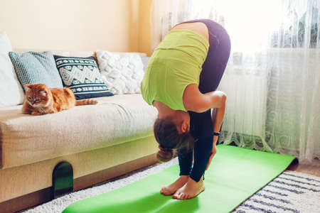 Home yoga workout during pandemic. Uttanasana standing forward bend pose. Woman training using mat by cat. Stretching exercisesの写真素材