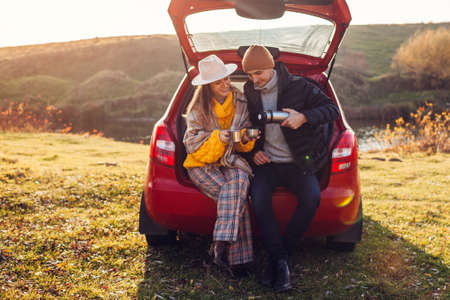 Loving couple relaxing in car trunk by autumn lake drinking tea from vacuum flask. Man and woman travel by auto. Young people enjoy fall activitiesの写真素材