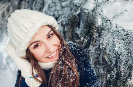 Close up portrait of young woman in winter park enjoying hoarfrost and snow by fir tree. Girl having fun outdoors wearing warm knitted hat and mittens.の写真素材