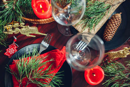Festive Christmas dinner table setting for two decorated with fir branches, red candles, cones, ornaments. Plates with cutlery and glasses surrounded with golden deerの写真素材