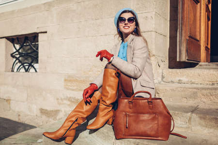Portrait of woman wearing stylish orange boots coat beret sitting on stairs by handbag outdoors. Fall female fashionable accesories. Vintage outfitの写真素材