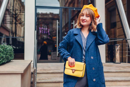 Portrait of stylish young woman wearing yellow beret blue coat holding purse outdoors. Spring fashion female accessories and clothes by modern architecture. Trendy classic retro outfitの写真素材