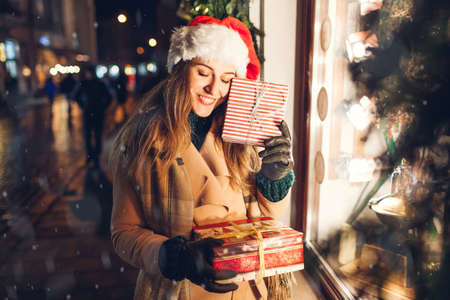 Happy woman in Santas hat holding presents gift boxes by shop showcase on city street at Christmas season.の写真素材