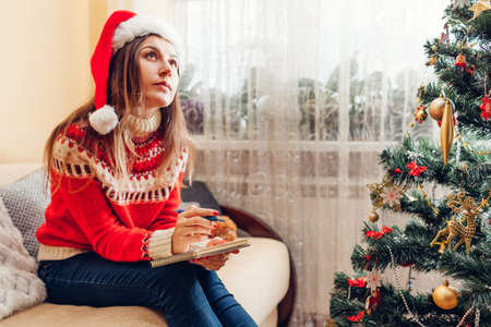 Young woman writing goals for 2022 New year in notebook at home sitting by Christmas tree in Santa...