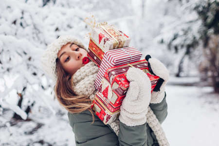 Young woman holding heap of heavy Christmas presents gift boxes in snowy winter park outdoors. Festive holiday season. Girl wearing warm knitted clothes and red lipstickの写真素材
