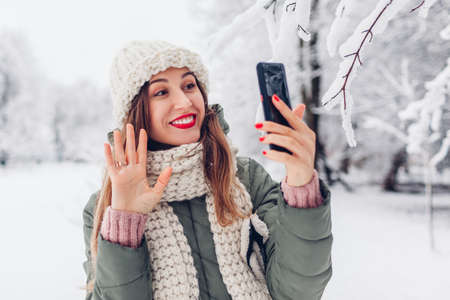 Portrait of young woman waving while calling on smartphone in snowy winter park through online video chat. Communication through internet in modern technologyの写真素材