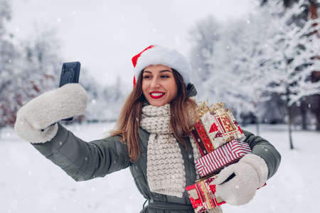 Portrait of young woman in Santa hat taking selfie on smartphone in snowy winter park holding heap of Christmas presents outdoors. Festive gift boxesの写真素材
