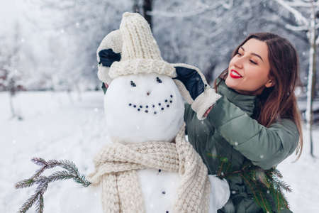 Young woman gives snowman outdoors in snowy winter park her hat, scarf and attaching fir branches in hands. Fun activities. Warm knitted clothes for friendの写真素材
