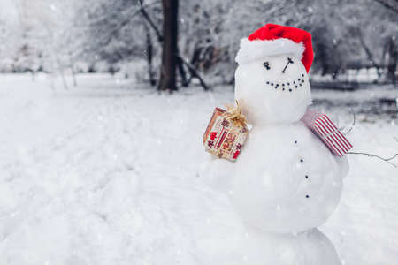 Happy snowman wearing festive Santa hat in snowy winter park at Christmas season outdoors holding presents gift boxes. Winter activitiesの写真素材