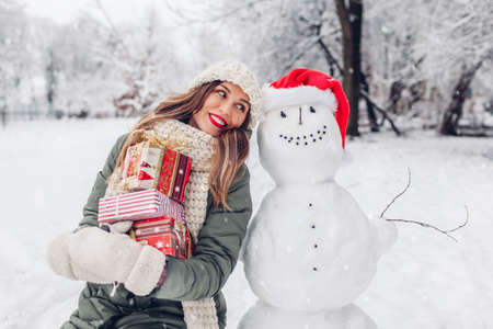 Happy woman holding heap of presents by snowman in Santa hat outdoors in snowy winter park. Festive Christmas holiday timeの写真素材