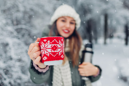 Woman drinking hot tea holding vacuum flask in snowy winter park. Cup dressed in red knitted...