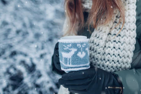 Close up of cup of tea or coffee dressed in knitted Christmas coat in female hand in winter park. Drinks to warm up in snowy frosty weather outdoorsの写真素材