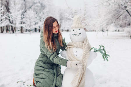 Girl makes snowman outdoors in snowy winter park dressing it in hat, scarf and attaching fir branches in hands. Seasonal fun activitiesの写真素材