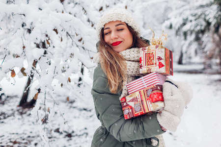Happy young woman holding heap of Christmas presents gift boxes in snowy winter park outdoors. Festive holiday season. Girl wearing warm knitted clothes and red lipstickの写真素材