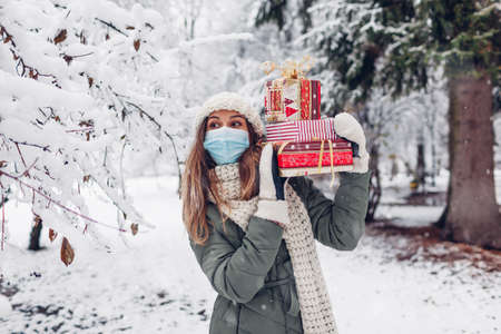 Young woman holding heap of Christmas presents gift boxes in snowy winter park outdoors wearing mask.の写真素材