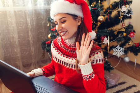Christmas lockdown. Woman in Santa hat waving hi in online video chat on laptop. Celebrating winter holidays at home.の写真素材