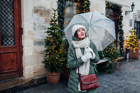Happy woman walking on city street by decorated for Christmas festive restaurant entrance holding umbrella under falling snow. Lviv during winter holidaysの写真素材