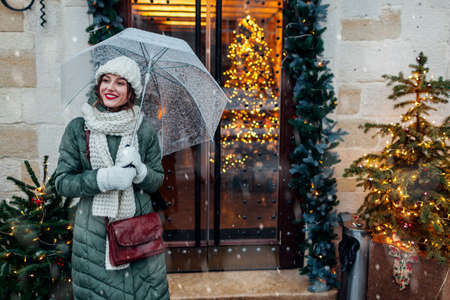 Happy young woman walking on city street by decorated for Christmas festive restaurant entrance holding umbrella under falling snow. Lviv during winter holidaysの写真素材