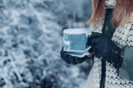 Close up of cup of tea or coffee dressed in knitted Christmas coat in female hand in winter park. Drinks to warm up in snowy frosty weather outdoorsの写真素材