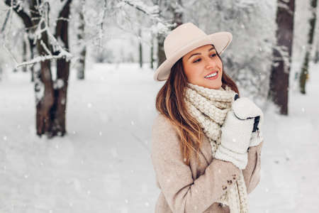 Portrait of stylish young woman in snowy winter park wearing fashionable white and beige warm hat coat scarf mittens. Young smiling girl walking outdoors. Female clothes and accessoriesの写真素材
