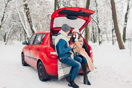 Loving couple sitting in car trunk drinking hot tea in snowy winter forest. People relaxing outdoors during road trip for holidaysの写真素材