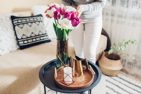 Woman enjoys tulips flowers put in vase on table with tray, watering can and candle. Cozy interior and spring decor at homeの写真素材
