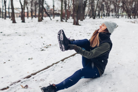 Runner woman fell down during training in snowy winter park. Woman feels pain in ankle lying on ground. Workout safetyの写真素材