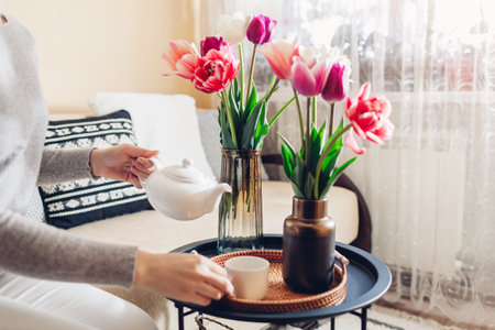 Woman pours tea out of kettle enjoying fresh tulips flowers in vases on coffee table. Interior and spring home decor.の写真素材