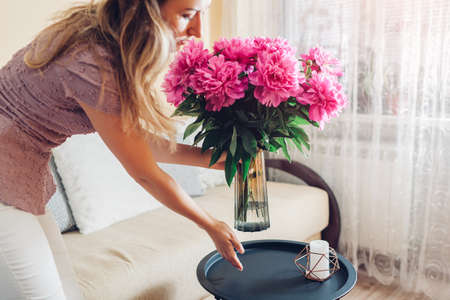 Woman puts vase with peonies flowers on table. Housewife taking care of coziness at home. Interior and spring decorの写真素材