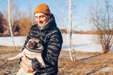 Young man walking pug dog in snowy winter park. Master playing with pet holding it on hands outdoors. Activities with animalsの写真素材