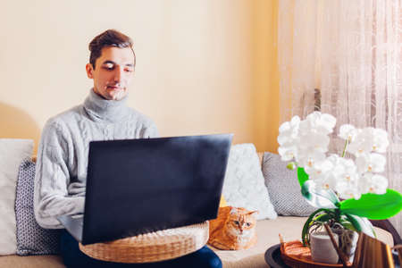 Man working online from home with pet using laptop sitting on couch. Freelancer typing on computer...