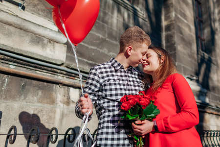 Valentines day couple in love. Man and woman hugging with bouquet of red roses flowers with balloons on city street. Holiday surprise giftの写真素材