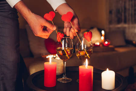 Man decorating table setting for Valentines day celebration at home putting hearts in glasses with champagne and strawberries on top by candles on table. Night dateの写真素材