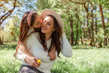 Girl kisses mother on cheek hugging while sitting on grass in spring park giving flower. Family relaxing outdoors. Mothers day. Spaceの写真素材