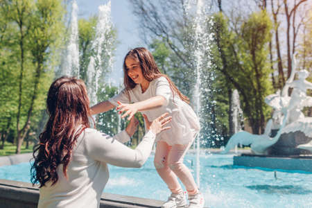 Young mother and her daughter having fun by fountain in spring park. Girl jumps in mom's hands. Mothers day. Family playing outdoorsの写真素材