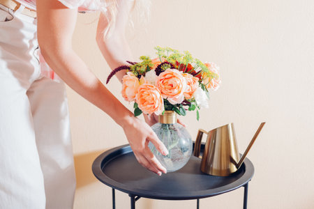 Woman puts vase with bouquet of flowers on table at home. Floral arrangement with orange roses. Interior and summer decorの写真素材