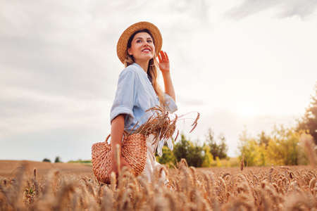 Portrait of happy young woman walking among wheat in summer field wearing straw hat holding handbag...