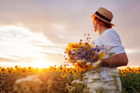 Senior woman holds bouquet of yellow sunflowers in summer field at sunset. Middle-aged woman admires landscapeの写真素材