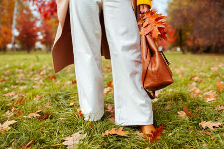Fashionable woman wearing stylish white flared trendy pants holding handbag and walking in fall park. Autumn female outfit in warm colors.の写真素材
