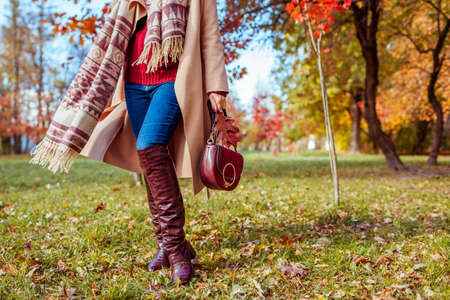 Fashionable woman wearing stylish clothes shoes and accessories walking in fall park. Autumn female outfit. Trendy burgundy knee high boots, coat and purse. Spaceの写真素材