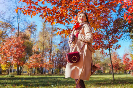 Happy young woman walking in autumn park among red trees. Female fall fashion. Stylish girl wearing beige coat and holding purseの写真素材