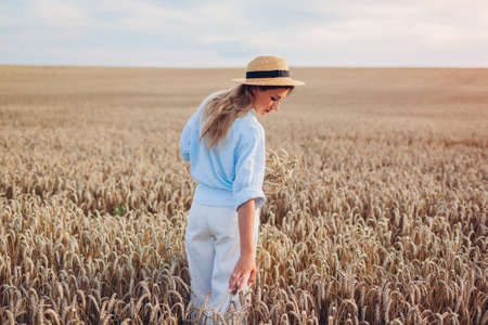 Young woman walking in summer field wearing straw hat and linen shirt holding wheat bundle. Girl touching wheat enjoying view at sunsetの写真素材