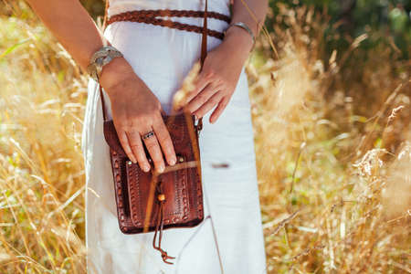 Close up of brown boho leather purse. Stylish woman wearing white dress, jewelry, holding handbag outdoors. Fashionable female accessoriesの写真素材