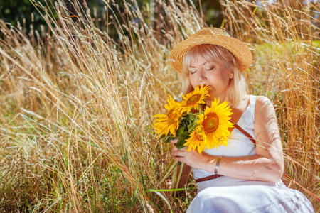 Middle-aged woman smells bouquet of yellow sunflowers relaxing in summer field with high grass in meadow outdoors. Harmony with natureの写真素材